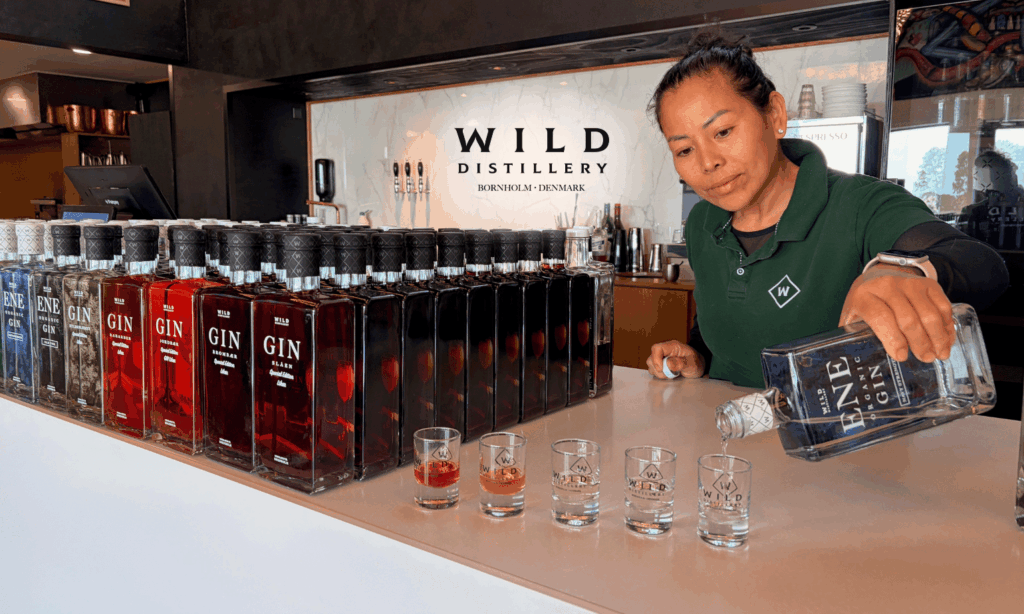 Sopha Nesrt ejer . grøn poloshirt skænker gin fra en Ene Organic Gin-flaske op i små smagsglas på en lys bardisk. Til venstre på disken står en lang, sirlig række af forskellige gin-flasker og likører i røde, mørke og klare nuancer. På marmorvæggen i baggrunden ses Wild Distillerys logo.