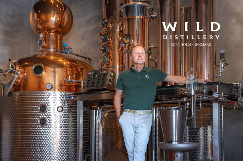 Anbefalet alt-tekst: Henrik Nerst fra Wild Distillery står foran det specialbyggede kobber-destilleringsapparat fra Müller Brennereianlagen til produktion af bornholmsk gin.